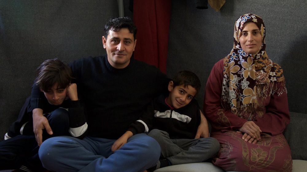 A Syrian man, Nemer, and his wife and two sons sit on pillows on a rug.